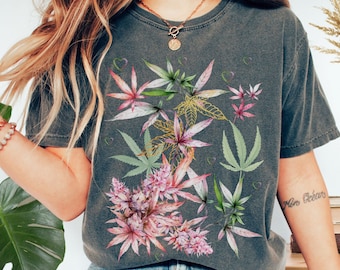 Può includere: Una maglietta grigia con una stampa floreale con foglie rosa e verdi. Le foglie sono stilizzate per assomigliare a piante di cannabis.