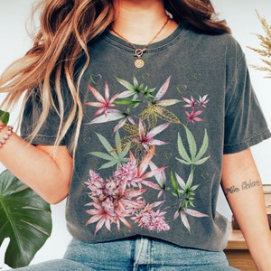 Puede incluir: Una camiseta gris con un estampado floral de hojas rosas y verdes. Las hojas están estilizadas para parecer plantas de cannabis.