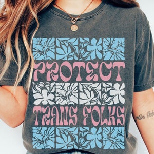 Może przedstawiać: Szary t-shirt z nadrukiem graficznym przedstawiającym stylizowany wzór kwiatowy w kolorze niebieskim i białym. Tekst "PROTECT TRANS FOLKS" jest nadrukowany na różowo i biało.