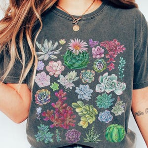 Puede incluir: Una camiseta gris oscuro con un estampado de suculentas coloridas. El estampado presenta una variedad de suculentas en tonos de verde, rosa, morado y rojo.