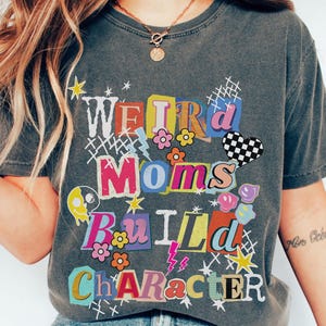 May include: Dark grey t-shirt featuring the text "WEIRD MOMS BUILD CHARACTER" in a colourful, collage-style design. The graphic includes flowers, stars, a chequered heart, and lightning bolts. The shirt is a casual, crew neck style.