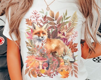 May include: White t-shirt featuring a watercolor illustration of a fox surrounded by autumn leaves, mushrooms, and foliage. The design includes shades of orange, brown, and green. The word "white" is printed at the bottom.
