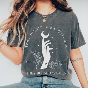 Pode incluir: T-shirt cinza escuro com o texto branco "THEY DIDN'T BURN WITCHES THEY BURNED WOMEN". O design inclui uma mão segurando uma lua crescente, com detalhes florais e celestiais. A camisa é combinada com jeans azuis e sapatos brancos.