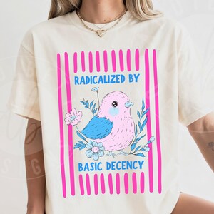 以下が含まれることがあります： ピンクの鳥と青い翼、花のアクセントが特徴のアイボリー色のTシャツ。ピンクの縦縞で縁取られています。「RADICALIZED BY BASIC DECENCY」のテキストが表示されています。Tシャツは半袖です。