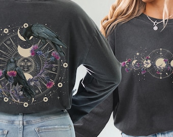 May include: Two long-sleeved shirts with a celestial design featuring moons, stars, and crows. One shirt has a single crow and moon design on the back, while the other shirt has a row of moons and crows across the front.