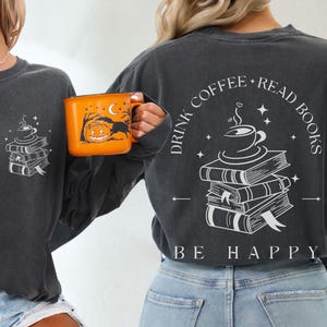 Op de afbeelding: Donkergrijze longsleeve met een witte afbeelding van een koffiekopje op boeken. De achterkant van de shirt heeft de tekst "DRINK COFFEE READ BOOKS BE HAPPY". Een oranje mok met een Halloween-ontwerp wordt vastgehouden.