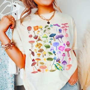 K&ouml;nnte beinhalten: Ein cremefarbenes T-Shirt mit einem bunten Pilz-Grafikdruck. Das Shirt hat einen Rundhalsausschnitt und kurze &Auml;rmel. Das Wort "Elfenbein" ist in Schwarz am unteren Rand des Bildes geschrieben.