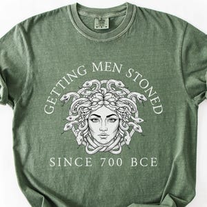 May include: Moss green t-shirt featuring a white graphic of Medusa with the text "GETTING MEN STONED SINCE 700 BCE". The shirt has a crew neck and short sleeves. The shirt is made of a soft, comfortable material.