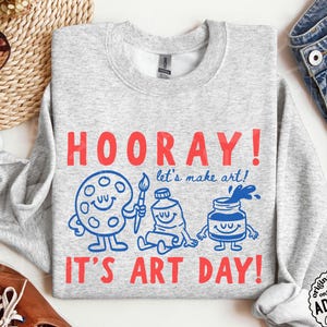 Könnte beinhalten: Ein hellgraues Sweatshirt mit Rundhalsausschnitt, rotem Text HOORAY! und IT'S ART DAY! sowie blauen grafischen Illustrationen einer lächelnden Farbpalette, einem Pinsel, einer Farbtube und einem Farbtopf mit dem blauen Text let's make art!.