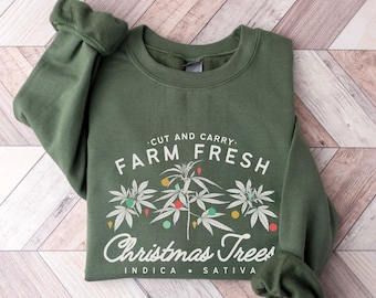 Può includere: Una felpa verde con la scritta "Cut and Carry. Farm Fresh Christmas Trees Indica Sativa" stampata sul davanti.