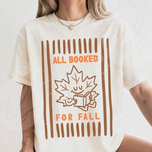 May include: An ivory short-sleeve t-shirt featuring a brown and orange graphic of a smiling maple leaf reading a book. The text reads ALL BOOKED FOR FALL, framed by vertical brown stripes. The design is centered on the front of the garment.