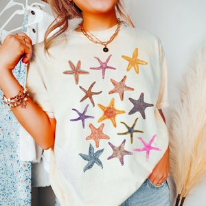 May include: Ivory-colored t-shirt featuring a collection of colorful starfish designs. The starfish are in various colors, including pink, yellow, blue, and purple. The word "ivory" is printed at the bottom of the image.