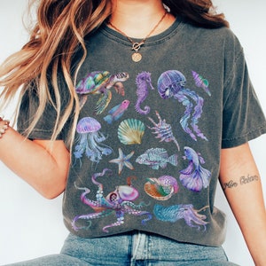 Könnte beinhalten: Ein dunkelgraues T-Shirt mit einem bunten Druck verschiedener Meerestiere, darunter eine Meeresschildkröte, Quallen, ein Seepferdchen, ein Oktopus und ein Seestern. Der Druck ist in Violett-, Blau- und Grüntönen gehalten.