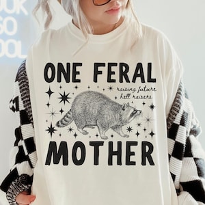 Op de afbeelding: Ivoor t-shirt met de tekst "ONE FERAL MOTHER" en een wasbeer illustratie. Het shirt bevat ook de zinnen "raising future" en "hell raisers" met ster accenten. Casual stijl.