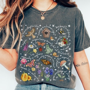 Op de afbeelding: Een donkergrijs t-shirt met een kleurrijk bloemen- en insectenontwerp, waaronder vlinders, bloemen en een paddenstoel. Het shirt heeft verschillende zinnen en woorden erop gedrukt, zoals "bee kind to all kinds" en "squash the patriarchy". Het woord "PEPPER" staat onderaan.