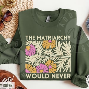 May include: An olive green crewneck sweatshirt featuring a graphic print of stylized cream-colored leaves and flowers in shades of pink and yellow. The text "THE MATRIARCHY" is printed above the floral design, and "WOULD NEVER" is printed below it.
