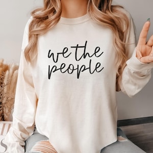 Könnte beinhalten: Elfenbeinfarbenes Langarmshirt mit dem Text "we the people" in schwarzer Schrift. Das Shirt hat eine neutrale Farbe und wird mit zerrissenen blauen Jeans kombiniert. Das Wort "IVORY" wird am unteren Bildrand angezeigt.
