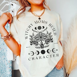 May include: A cream colored t-shirt with a black graphic design that says "Witchy Moms Build Character" with a moth, moon, and stars.