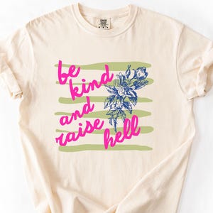 May include: Ivory-colored t-shirt with the text "be kind and raise hell" in pink, along with a floral design in blue and green. The shirt has a crew neck and short sleeves.