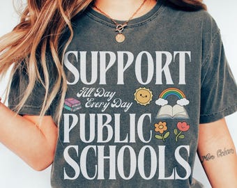 Könnte beinhalten: Dunkelgraues T-Shirt mit den weißen Worten "SUPPORT PUBLIC SCHOOLS". Das Shirt zeigt eine Sonne, einen Regenbogen, ein Buch und eine Blumengrafik. Das Wort "Pepper" steht unten.