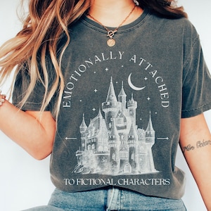 May include: A dark gray t-shirt with a white graphic of a castle, stars, and a crescent moon. The text "Emotionally Attached to Fictional Characters" is printed on the shirt.
