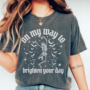 May include: Dark gray t-shirt with the text "on my way to brighten your day" and a dancing skeleton graphic. The design includes bats, stars, and a crescent moon. The shirt is a casual style.