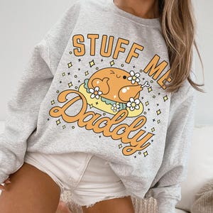 Pu&ograve; includere: Una felpa grigio chiaro con la scritta "STUFF ME Daddy" in lettere arancioni. La grafica presenta un tacchino dei cartoni animati con fiori. La felpa &egrave; uno stile girocollo.