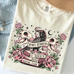 Könnte beinhalten: Elfenbeinfarbenes T-Shirt mit einem Grafikdesign mit einem Raben, Totenköpfen, Rosen und dem Text "REBEL READERS", "BANNED BOOK" und "SOCIAL CLUB". Das Shirt wird mit einer Jeans und einer Halskette präsentiert.