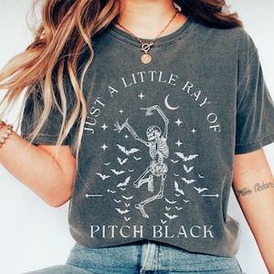 Pode incluir: Uma camiseta cinza escura com um gráfico branco de um esqueleto dançando com morcegos e uma lua crescente. O texto "Just a little ray of pitch black" está impresso na camiseta.