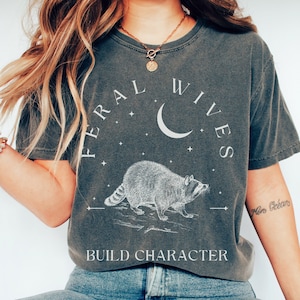 Pode incluir: Camiseta cinza escuro com o texto "FERAL WIVES" acima de uma ilustração de guaxinim e uma lua crescente. A frase "BUILD CHARACTER" é impressa abaixo. A camisa é de estilo vintage.