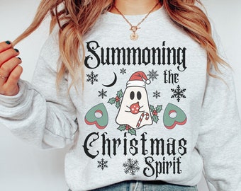 Op de afbeelding: Een grijs sweatshirt met een wit grafisch ontwerp van een geest die een kerstmanmuts draagt en een snoepstok vasthoudt. De tekst "Summoning the Christmas Spirit" is in zwart op het sweatshirt gedrukt.