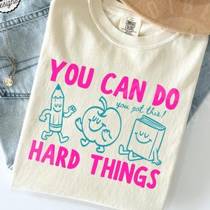 May include: Ivory t-shirt with the text "YOU CAN DO HARD THINGS" in pink. The shirt features cartoon illustrations of a pencil, apple, and book. The shirt is part of the Comfort Colors collection.