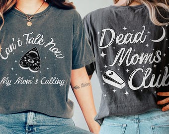 May include: Dark gray t-shirts with white text and graphics. One shirt reads "Can't Talk Now, My Mom's Calling" with a planchette design. The other shirt says "Dead Moms Club" with a coffin and moon.