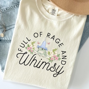 以下が含まれることがあります： 黒いスクリプトで「FULL OF RAGE AND Whimsy」というフレーズが書かれたアイボリー色のTシャツ。青い帽子をかぶったカエルのイラストが特徴で、花と蝶に囲まれています。Tシャツはブルージーンズとネックレスと組み合わせています。