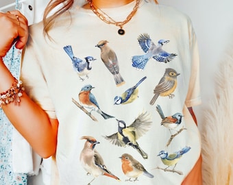 May include: A light yellow t-shirt with a colorful print of various birds. The birds are depicted in a watercolor style and include bluebirds, robins, and sparrows. The shirt is a relaxed fit and has a crew neck.