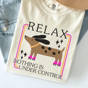 May include: Ivory-colored t-shirt with the words "RELAX" and "NOTHING IS UNDER CONTROL" in black. A cartoon dog with black ears and spots, wearing pink boots, is in the center. The shirt is folded on top of blue jeans and a tan hat.