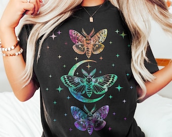 May include: Black t-shirt with a graphic of three iridescent moths and a crescent moon with stars. The moths are in a vertical line, with the top moth being the largest and the bottom moth being the smallest. The crescent moon is in the middle of the graphic, and the stars are scattered around the moths and moon.