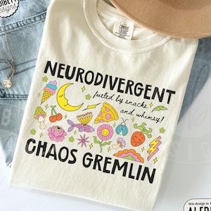 Könnte beinhalten: Ein elfenbeinfarbenes T-Shirt mit dem schwarzen Text NEURODIVERGENT fueled by snacks and whimsy! CHAOS GREMLIN. Die Grafik enthält bunte Illustrationen einer Eistüte, einer Mondsichel, eines Pizzastücks, eines Pilzes, Kirschen, eines Fisches, einer Blume, eines Marienkäfers und eines Blitzes.