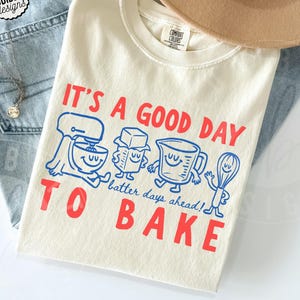 Könnte beinhalten: Ein elfenbeinfarbenes T-Shirt mit rotem Text, der IT'S A GOOD DAY TO BAKE lautet, und blauem Text, der batter days ahead! lautet. Die Grafik enthält blaue Cartoon-Illustrationen einer Küchenmaschine, eines Stücks Butter, eines Messbechers und eines Schneebesens.
