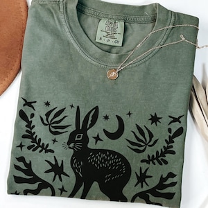 以下が含まれることがあります： モスグリーンのTシャツ。黒いウサギ、星、葉のグラフィック。クルーネックと半袖。金のネックレスがTシャツの上にかけられています。「Comfort Colors」と書かれたタグが付いています。