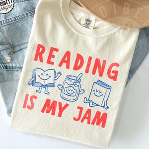 May include: An ivory t-shirt featuring the red text READING IS MY JAM. Between the lines of text are blue line-art illustrations of a smiling open book, a smiling jar of jam with a spoon, and a smiling closed book sitting on the fabric.
