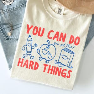 Può includere: T-shirt color avorio con la scritta "YOU CAN DO HARD THINGS" in rosso e blu. La maglietta presenta illustrazioni di cartoni animati di una matita, una mela e un libro. La maglietta è di Comfort Colors.