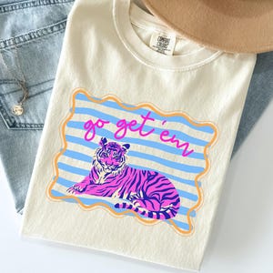 Pu&ograve; includere: T-shirt color avorio con grafica di tigre rosa e blu e la scritta "go get 'em". Il design &egrave; posto su strisce blu e un bordo arancione. La maglietta &egrave; abbinata a jeans blu e una collana.