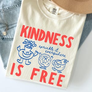 Puede incluir: Camiseta de color marfil con las palabras "KINDNESS IS FREE" en rojo, junto con ilustraciones de dibujos animados azules. La camiseta está doblada sobre unos vaqueros azules, un collar y un sombrero beige.