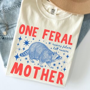 May include: Ivory-colored t-shirt with the words "ONE FERAL MOTHER" in red, and a blue raccoon illustration. The shirt also has the text "raising future hell raisers" in blue. The shirt is folded on top of blue jeans.