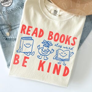 May include: An ivory t-shirt featuring the red text READ BOOKS BE KIND and the blue text stay weird. The graphic includes three cartoon characters: a book with a smiling face, a flower vase with a smiling face, and an open book with a smiling face.