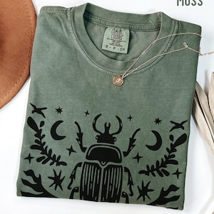 May include: Moss green t-shirt with a black beetle and celestial design. The shirt has a crew neck and short sleeves. A gold necklace with a pendant is draped across the shirt. The tag reads "Comfort Colors".