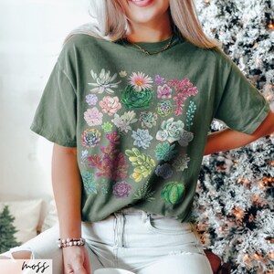 Puede incluir: Una camiseta verde con un estampado de suculentas coloridas. La camiseta tiene un corte holgado y cuello redondo.