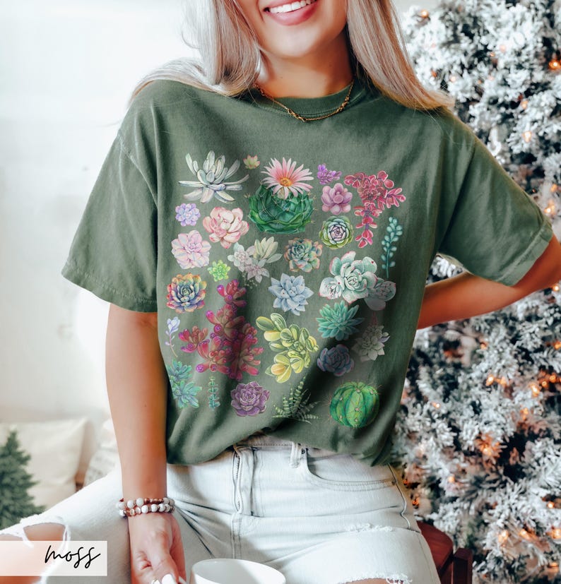 Puede incluir: Una camiseta verde con un estampado de suculentas coloridas. La camiseta tiene un corte holgado y cuello redondo.
