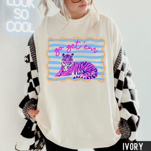 Pu&ograve; includere: T-shirt color avorio con grafica di una tigre viola a strisce blu, su sfondo a righe blu e arancioni. Il testo "go get 'em" &egrave; scritto sopra la tigre in rosa. La maglietta ha una vestibilit&agrave; ampia.